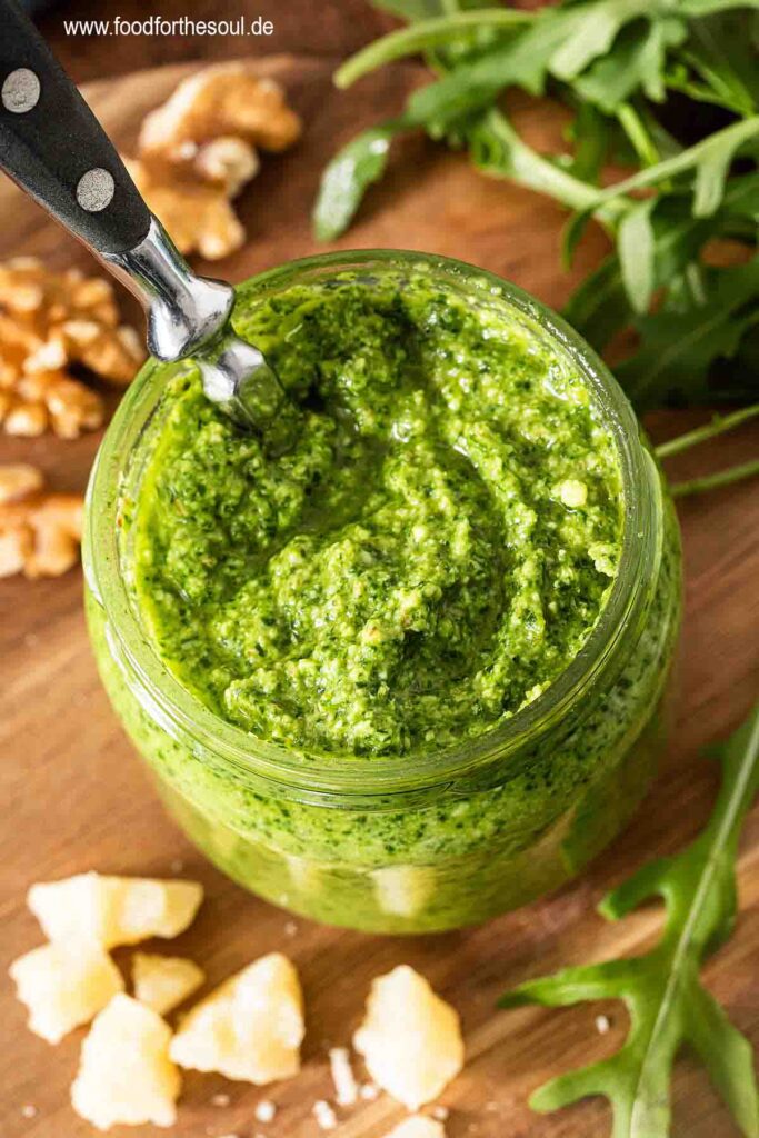Selbstgemachtes Rucola Pesto in einem Glas mit Löffel umgeben von Zutaten