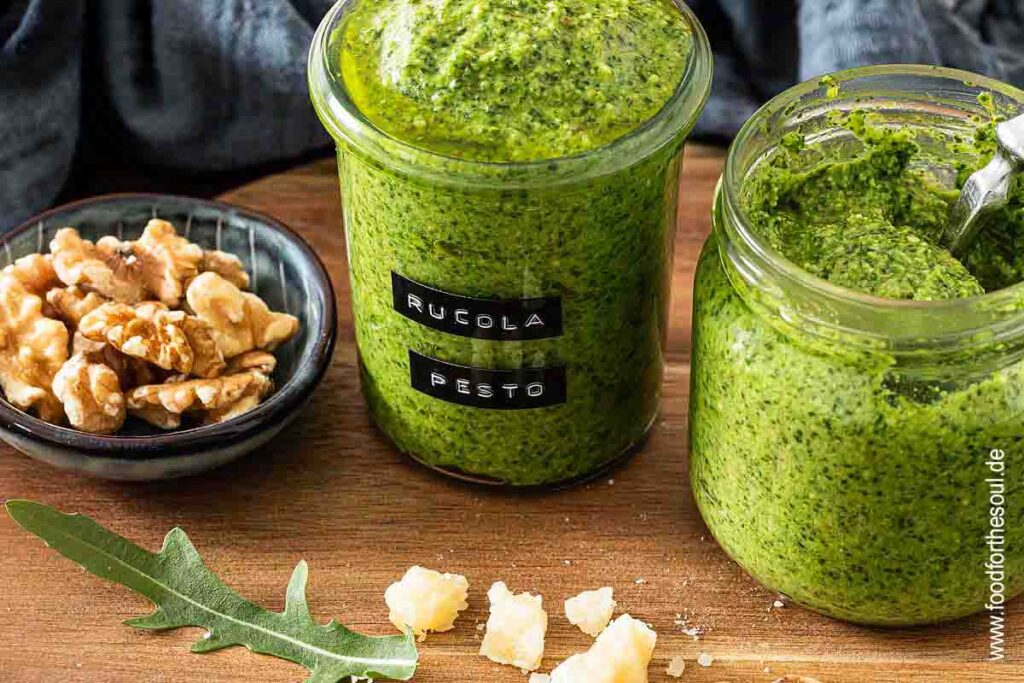 Selbstgemachtes Rucola Pesto in einem beschrifteten Glas, daneben ein weiteres Glas Pesto und eine Schale mit Walnüssen