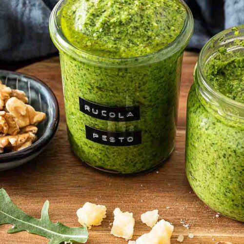 Selbstgemachtes Rucola Pesto in einem beschrifteten Glas, daneben ein weiteres Glas Pesto und eine Schale mit Walnüssen