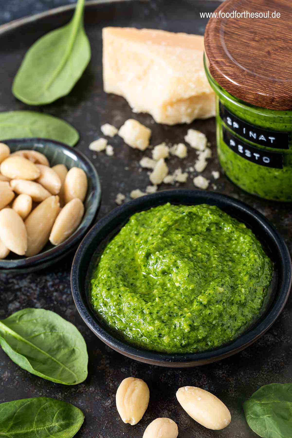 Frisch zubereitetes Spinat Pesto in einer Schale, daneben Mandeln, Parmesan und Spinatblätter